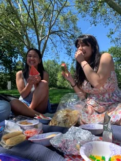 two women sitting on a blanket eating food and drinking watermelon slices in the park