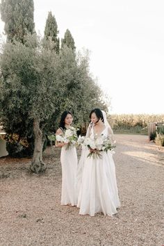 two women in white dresses standing next to each other near an olive tree and bushes