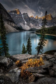 a lake surrounded by trees and rocks under a cloudy sky with mountains in the background