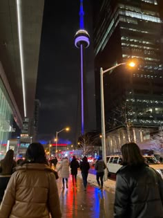 people are walking down the sidewalk in front of tall buildings at night with lights on