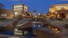 people are walking around in front of shops and water features at dusk, with the lights on