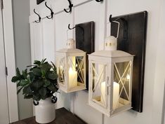 two white lanterns are hanging on the wall next to a potted plant and candle holders