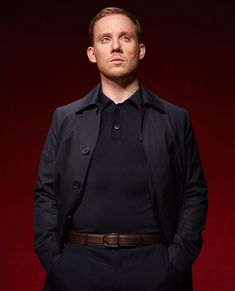 a man in a black shirt and suit standing with his hands in his pockets while wearing a brown belt