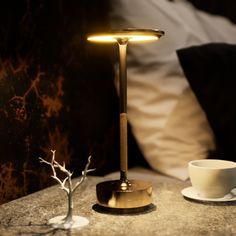 a table with a lamp on it next to a cup and saucer in front of a bed