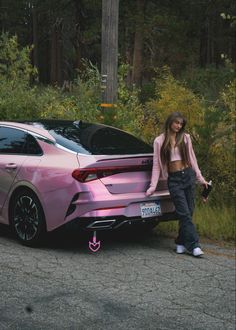 a woman standing next to a pink car on the side of the road with trees in the background