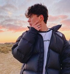 a young man in a puffy jacket standing on the beach at sunset with his hand to his face