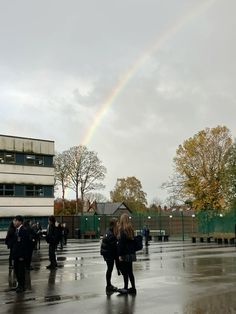 two people are standing in the rain under a rainbow over an open area with buildings and trees