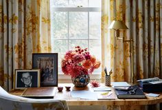 a desk with flowers and pictures on it in front of a window covered by drapes