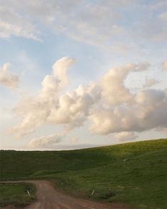 a dirt road in the middle of a green field with white clouds above it on a sunny day