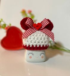 a small white and red doll with a bow on it's head sitting next to a heart