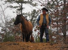 a man standing next to a brown horse on top of a dirt hill with trees in the background