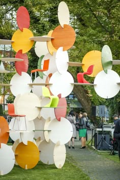 many different colored circles hanging from a wooden structure in the grass with people walking by