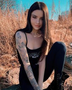 a woman sitting on top of a rock next to dry grass and bushes with tattoos on her arms