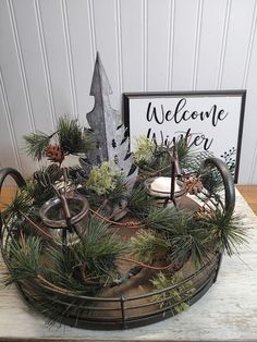 a welcome winter sign with pine cones and evergreen branches in a metal basket on a wooden table