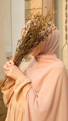 a woman wearing a pink hijab and holding dried flowers in front of her face