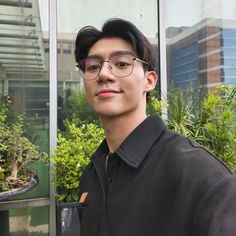 a young man wearing glasses standing in front of a bonsai tree and potted plants