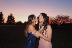 two young women are kissing each other in front of the setting sun on a field