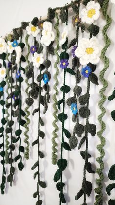 crocheted flowers are hanging on the wall next to green leaves and stems with blue, white, and yellow centers