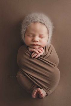 a newborn baby wrapped in a brown wrap and wearing a grey hat with his hands under his chin