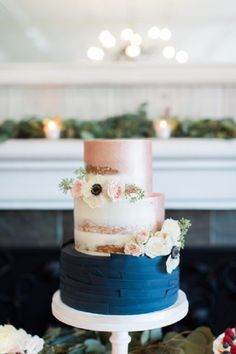 a three tiered cake with pink and blue frosting on top sits on a table in front of a fireplace