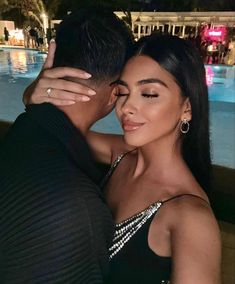 a man and woman embracing in front of a swimming pool at night with lights on