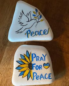 two white painted rocks with peace and pray for peace written on the top one has a sunflower