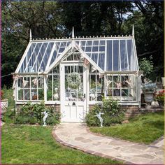 a small white greenhouse in the middle of a garden with lots of flowers and greenery