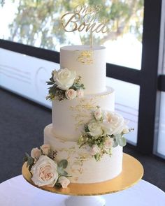 a three tiered wedding cake with white flowers and gold trimmings on a table