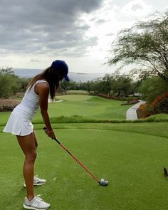 a woman in white dress playing golf on green field with trees and cloudy sky behind her