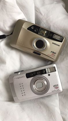 two cameras sitting next to each other on top of a white bed sheet, one is silver and the other is black