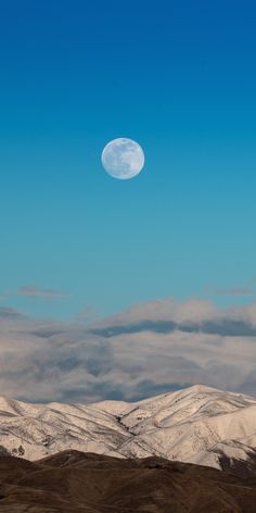 the moon is shining in the blue sky above some hills and snow - capped mountains