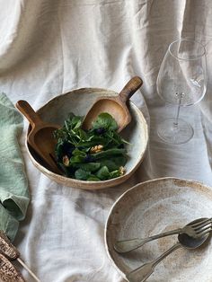 a bowl filled with greens next to two wine glasses and spoons on a white table cloth