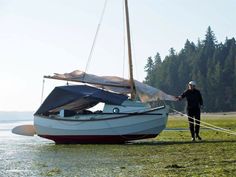 a man standing next to a sailboat on the shore with trees in the background