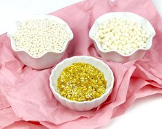 three white bowls filled with yellow and white sprinkles on top of pink paper