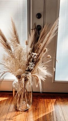 dried flowers in a glass vase sitting on the floor next to a door with blinds