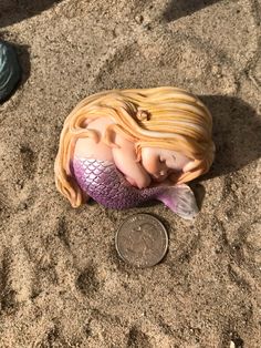 a mermaid doll laying on the sand next to a penny