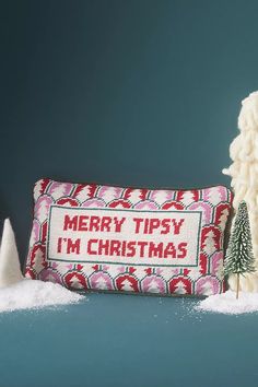 a cross - stitch christmas pillow sitting next to a small pine tree on a snowy surface