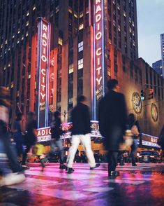 people walking in the city at night with buildings and signs lit up behind them that read radio city