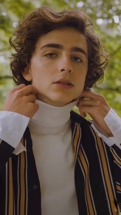 a young man with curly hair wearing a white turtle neck sweater and black striped jacket