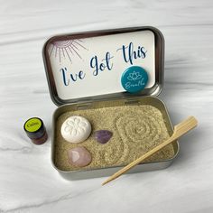 an open tin with sand and rocks in it next to a wooden stick on the ground