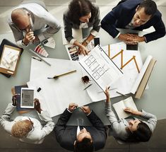 four people sitting at a table with papers and pens in front of them, looking down