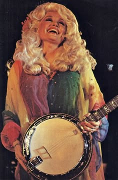 a woman with long blonde hair holding a banjo in her right hand and smiling at the camera