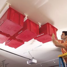 a man standing on a ladder painting the ceiling with red boxes hanging from it's ceiling