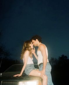 a man and woman sitting on the hood of a car in front of a dark sky