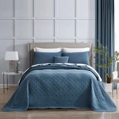 a bed with blue bedspread and pillows in a white room next to a window