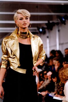 a woman in a gold jacket and black dress on the catwalk at a fashion show