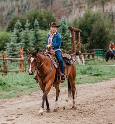 a woman riding on the back of a brown horse down a dirt road next to a forest