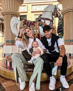 a man and woman sitting on a bench with a baby in front of a carousel