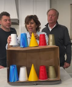 two men and a woman standing around a cardboard box with cups on it