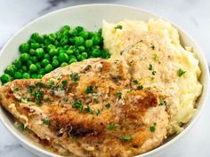 a white bowl filled with mashed potatoes and green peas next to meat on the side